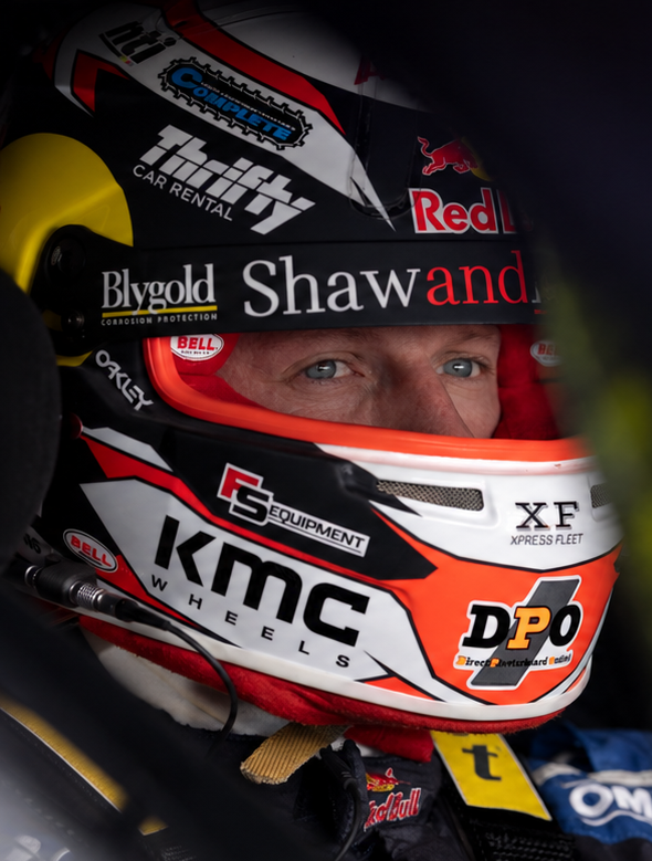 A close-up lifestyle shot of a race driver wearing a KMC Wheels DPO #01 racing helmet with various sponsor logos, inside a race car cockpit.